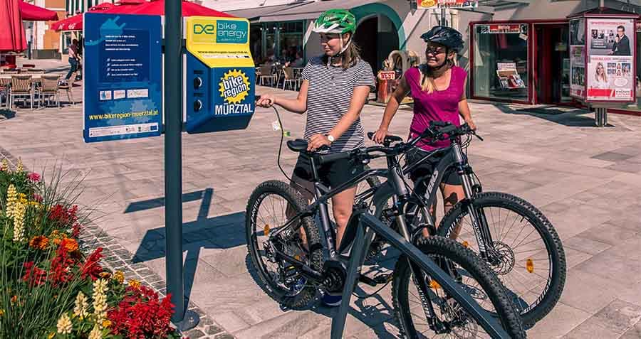 zwei-junge-frauen-laden-ihr-e-bike-an-der-ladestation-auf-in-der-bike-region-muerztal